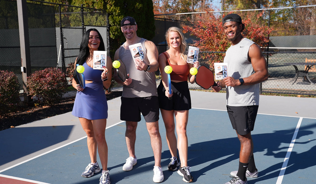 A group of four pickleball players on an outdoor court holding Pickle Putty and paddles, showing how the no-bend ball pickup tool works.